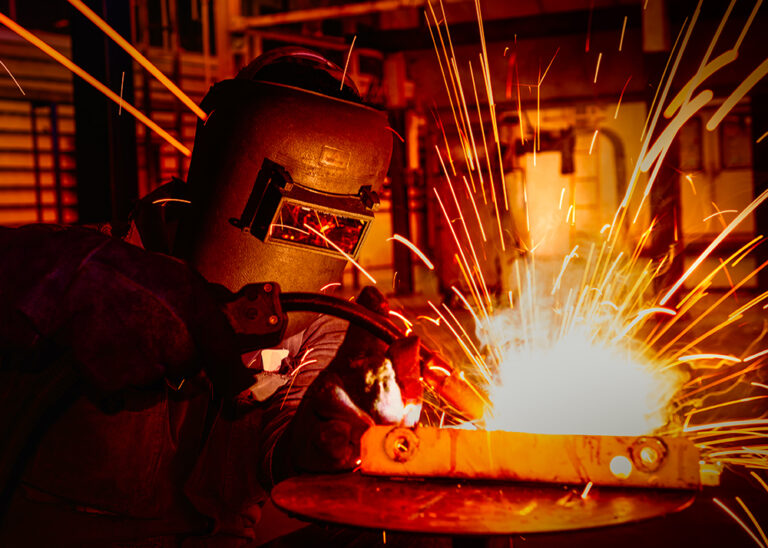 Welders in protective gear with a welding torch on metal, with bright sparks flying around in a dimly lit workshop.